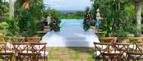 Pasillo nupcial al aire libre con sillas y decoración floral, que conduce a una piscina.
