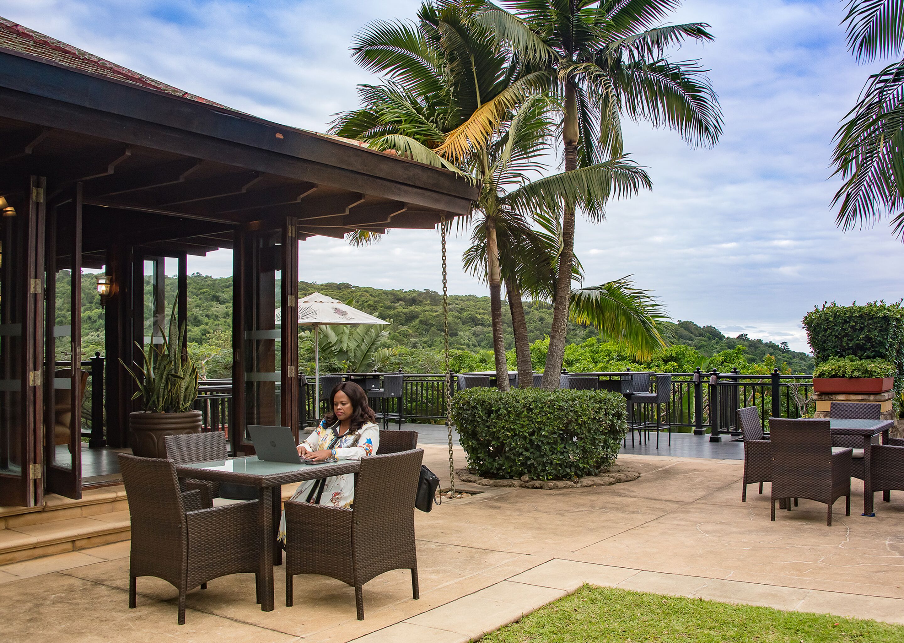 An outdoor patio with wicker furniture, a woman working on a laptop, and a view of lush greenery and hills in the background.