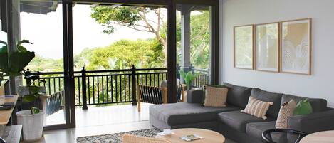 A modern living room with a grey sofa, a round wooden coffee table, a wicker chair, and a balcony with a view of greenery.