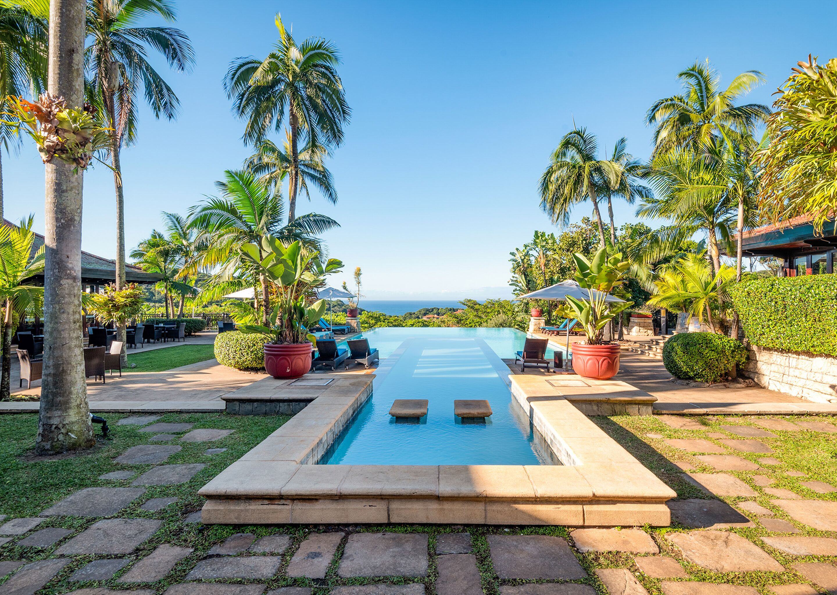 Ein Poolbereich mit Blick aufs Meer, umgeben von Palmen und Liegestühlen.