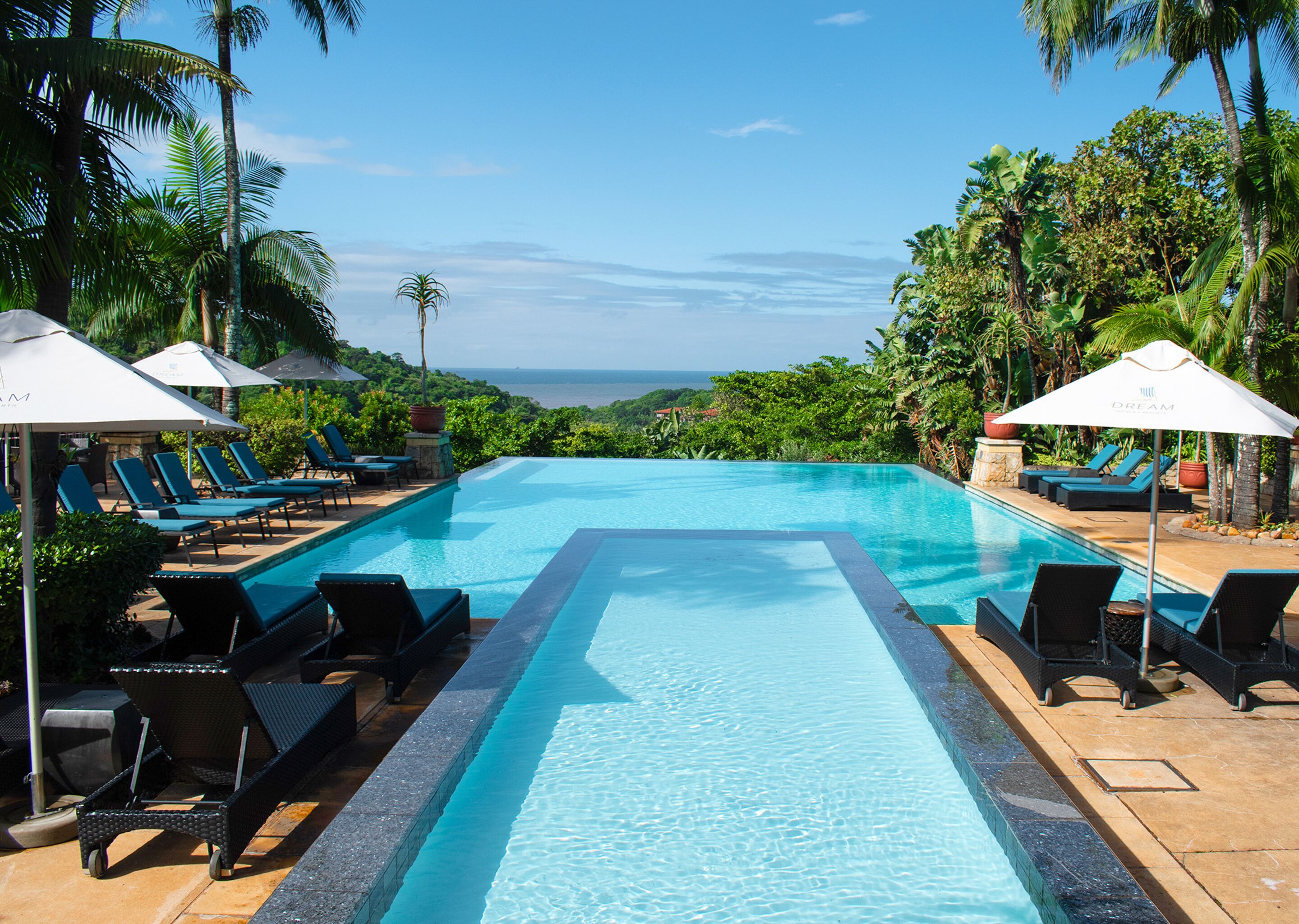 A pool with lounge chairs and umbrellas, surrounded by palm trees and a view of the ocean.