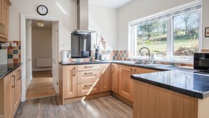 A modern kitchen with wooden cabinets, a black countertop, and a large window providing a view of the outdoors. - Sorrento - 3 Bed Holiday Home - Wisemans Bridge (Narberth)