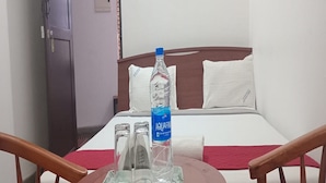 A hotel room with a bed, a bedside table with a water bottle and glasses, and a wooden chair. - Mehaa Paradise Family Guest House (Puducherry)