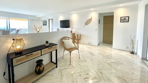 A modern living room with a console table, a wicker chair, a flat-screen TV mounted on the wall, and a large window offering a view of the city. - Vezelay 3 (Cannes)