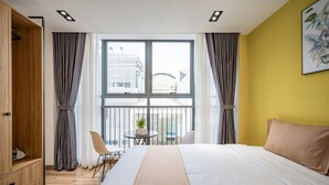 Habitación de hotel con una cama grande, un armario de madera, una mesita con una planta, una silla y una ventana con cortinas.