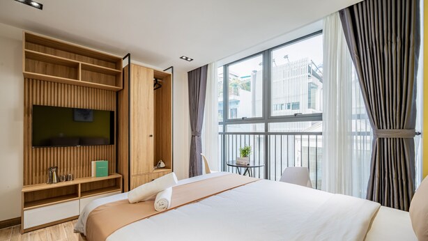 Habitación de hotel con cama, televisor montado en la pared, una mesita con una planta y una ventana con cortinas.