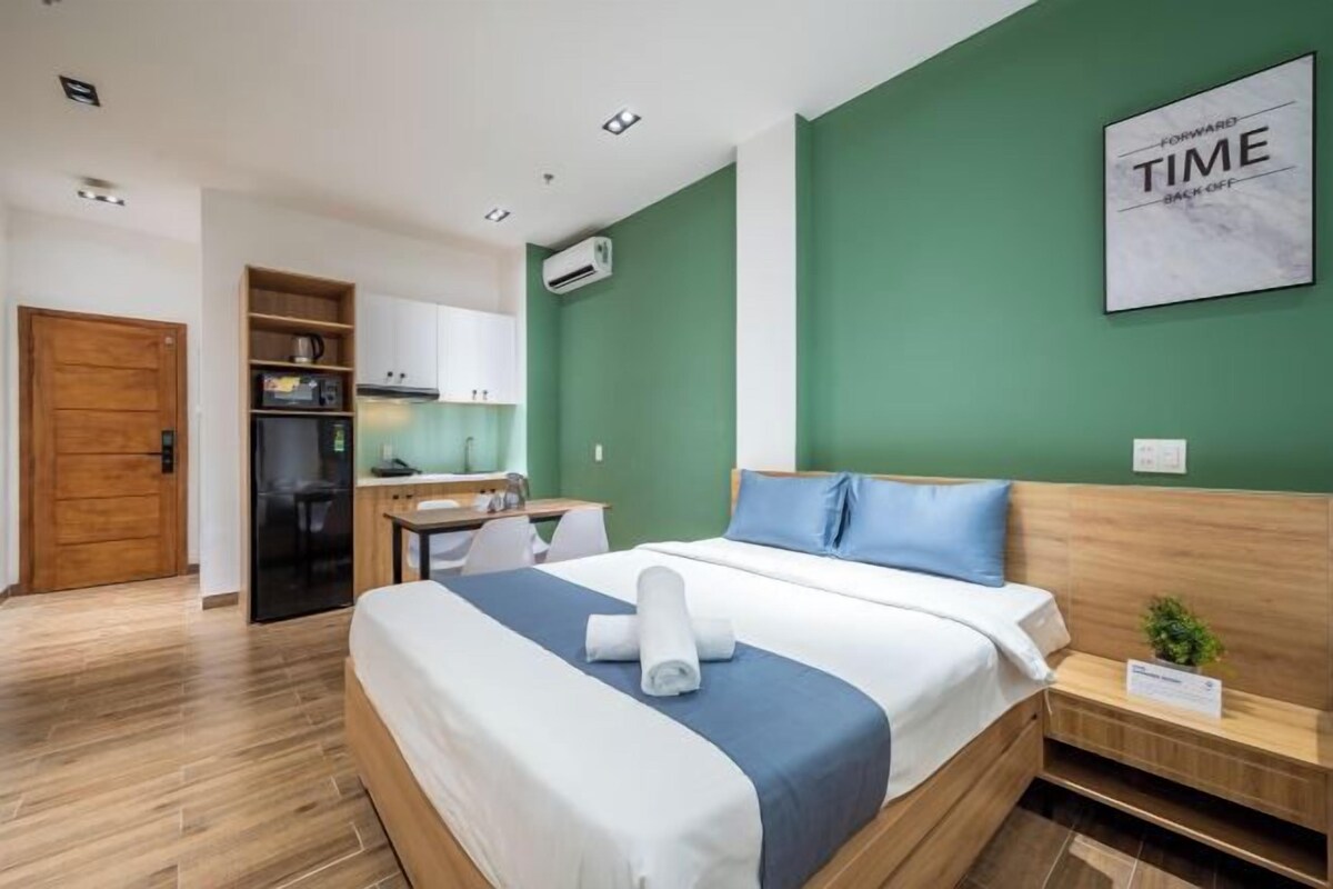 Quarto de hotel com cama, escrivaninha, cadeira, mesa pequena, planta e parede verde com frase motivacional.
