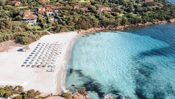 Vista aérea de una zona costera con playa, aguas turquesas cristalinas y un conjunto de casas rodeadas de vegetación.