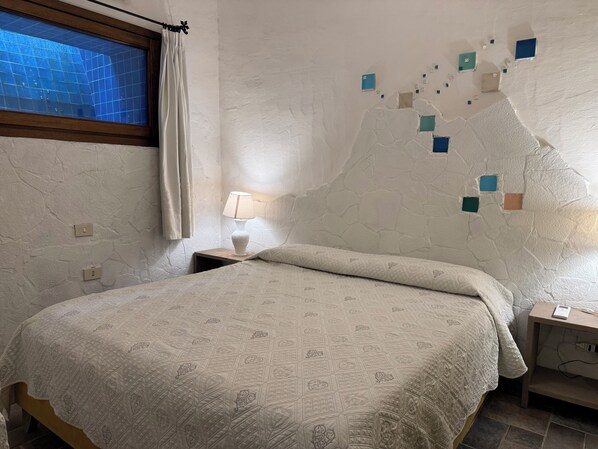 A bedroom with a bed, a nightstand with a lamp, a wall with a textured pattern, and a window with a curtain. - Villa Astra Fronte Mare - Porto Cervo (Porto Cervo)