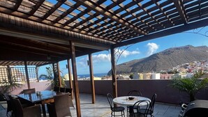 An outdoor patio with a pergola, a dining table with chairs, a grill, and a view of a city and mountains in the background. - Gomera Isla Mágica (San Sebastián de La Gomera)