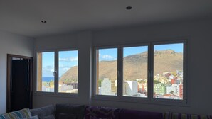 A living room with a sofa and a coffee table, large windows with a city and mountain view, and a door. - Gomera Isla Mágica (San Sebastián de La Gomera)
