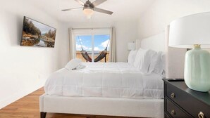 A bedroom with a bed, a ceiling fan, a hammock, a window with a view, and a nightstand with a lamp.