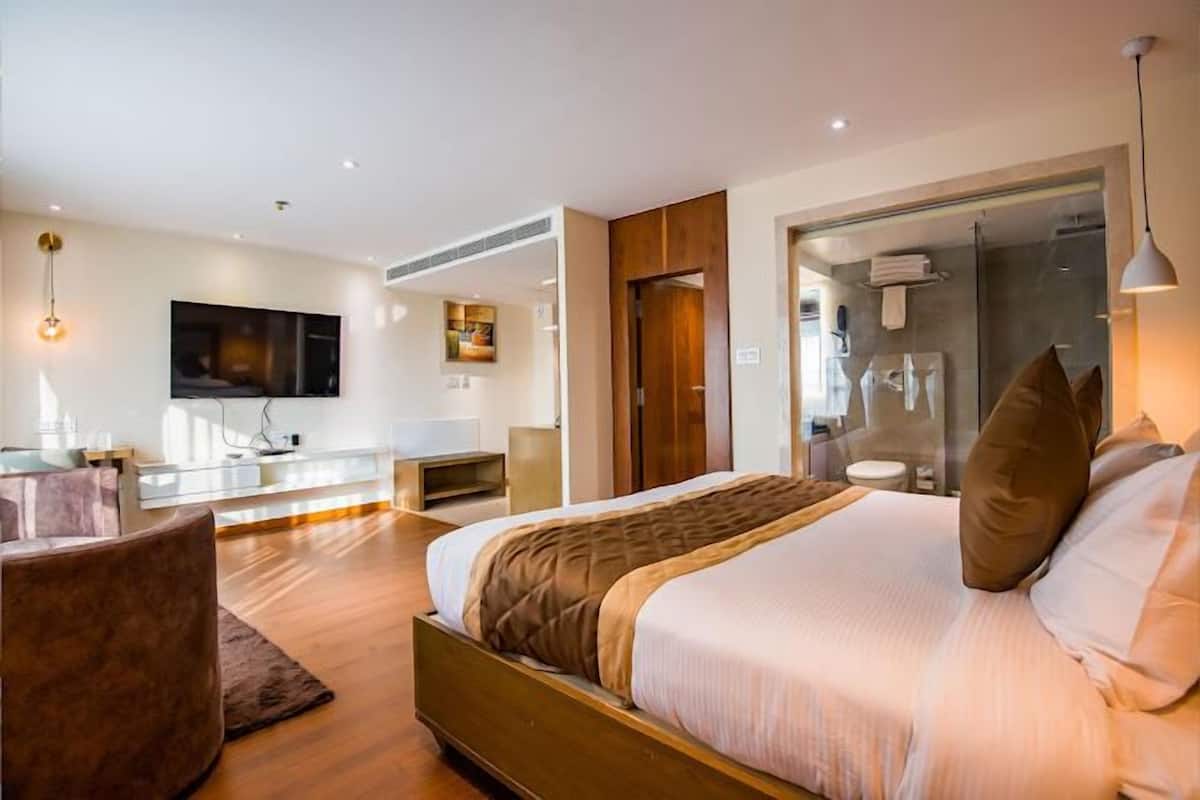 A modern hotel room with a large bed, a seating area, a TV, and a glass shower enclosure.