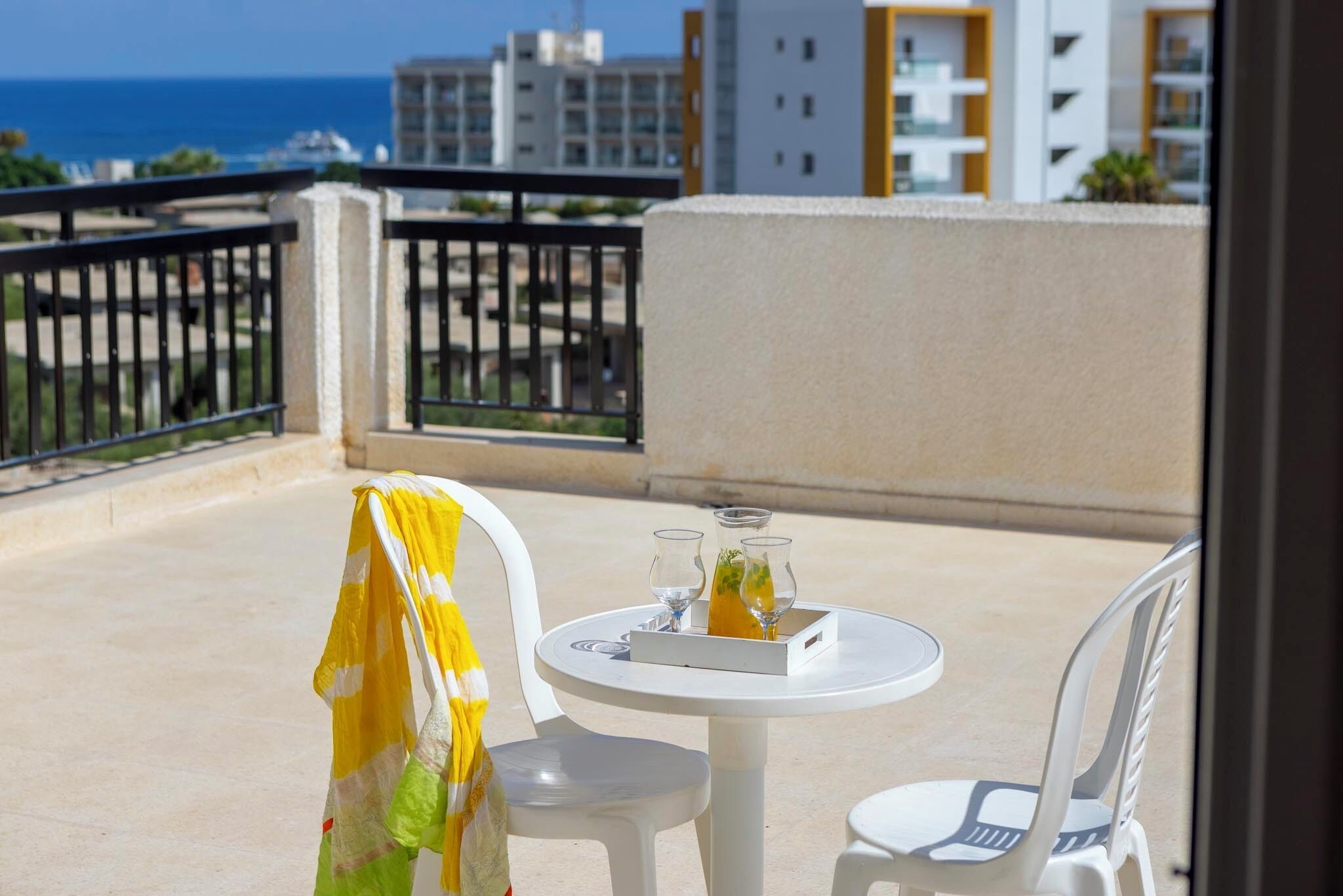 A balcony with a table and chairs, a drink tray, and a view of the sea and buildings.
