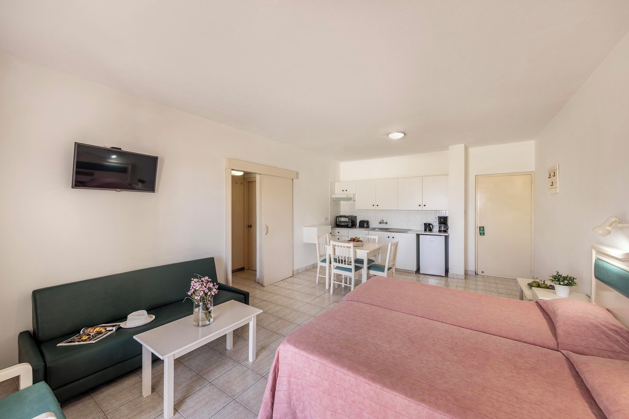 A compact hotel room with a bed, a sofa, a small table, a TV, a kitchenette, and a bathroom.
