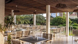 Un patio al aire libre con mesas y sillas blancas, techo de paja y follaje al fondo.