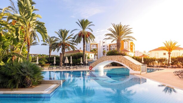 Una piscina con un puente de piedra, palmeras y sillones de descanso.