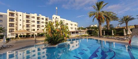 A swimming pool with a clear blue water surface, surrounded by palm trees and a white building with balconies.