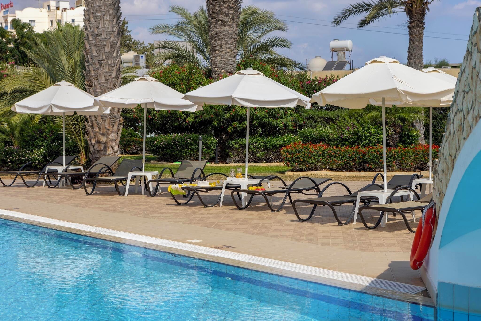 Zona de piscina con sillones de descanso y sombrillas, rodeada de palmeras y una piscina azul cristalina.
