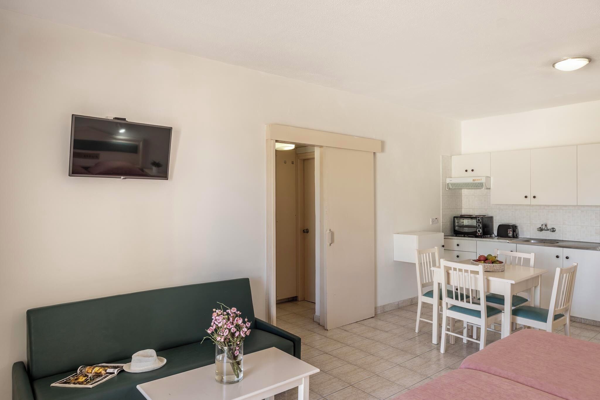 A room with a green sofa, a small table with a vase of flowers, a TV mounted on the wall, and a kitchenette area with a microwave, toaster, and cabinets.