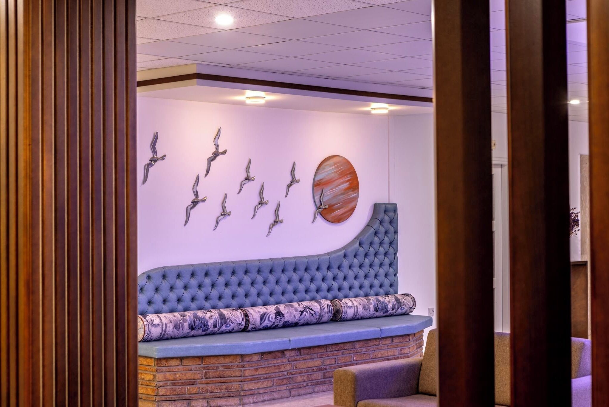 A modern lounge area with a tufted blue sofa, a brick accent wall, and a wall-mounted sculpture of birds.