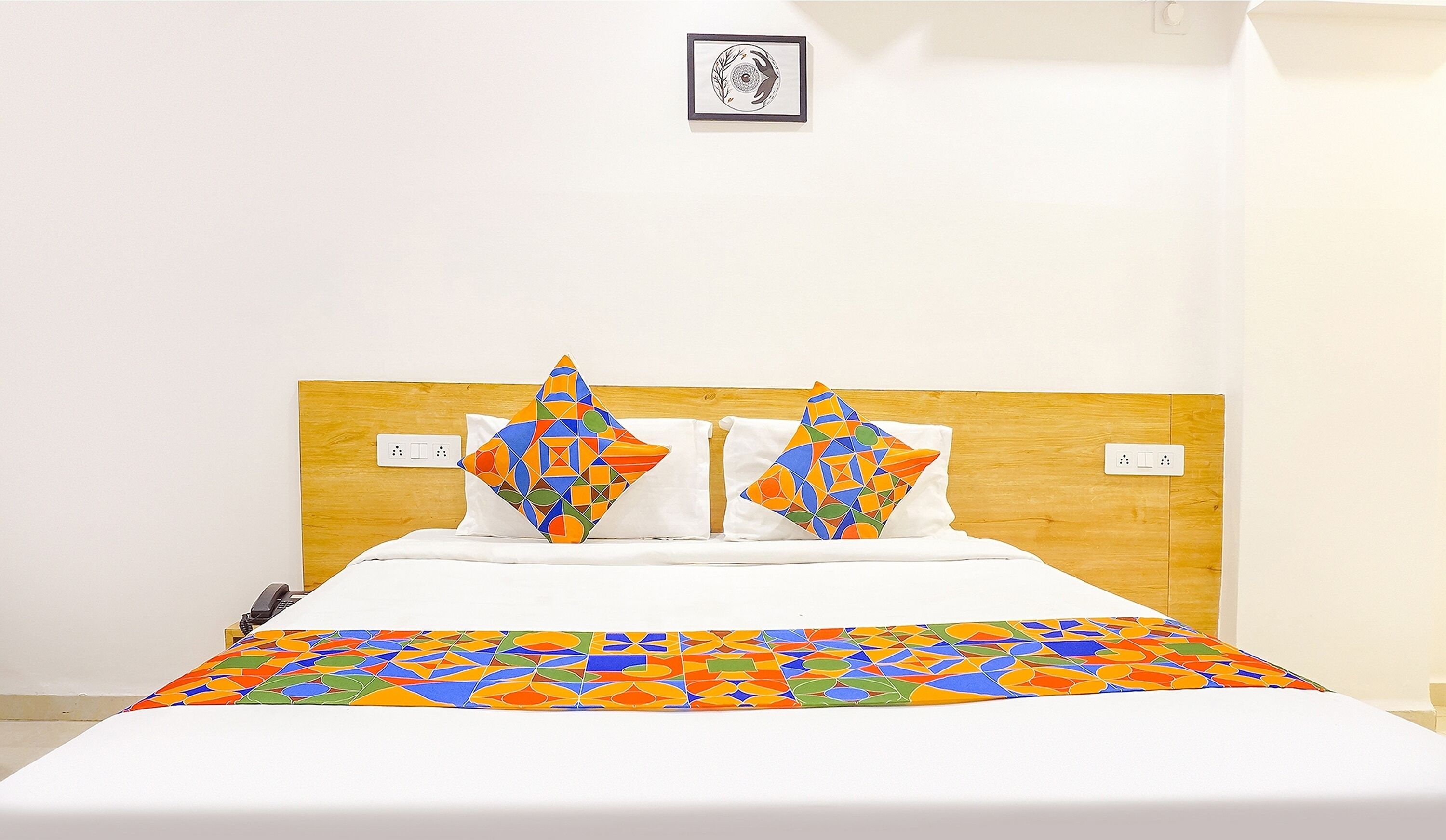 A bed with a wooden headboard, two decorative pillows, a colorful bedspread, and a small framed picture on the wall.