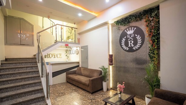 A hotel lobby with a staircase, a reception desk, a brown sofa, a coffee table, and a decorative wall with a crest. - Hotel Royce Executive (Mumbai)