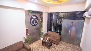 A lobby with a sofa, a coffee table, a potted plant, and a wall with a logo. - Hotel Royce Executive (Mumbai)