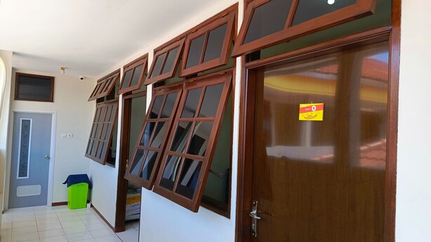 A hallway with a wooden door, a green trash bin, and multiple windows with wooden frames. - Bebek Kuning Residence (Malang)