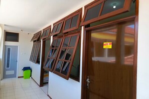 A hallway with a wooden door, a green trash bin, and multiple windows with wooden frames. - Bebek Kuning Residence (Malang)