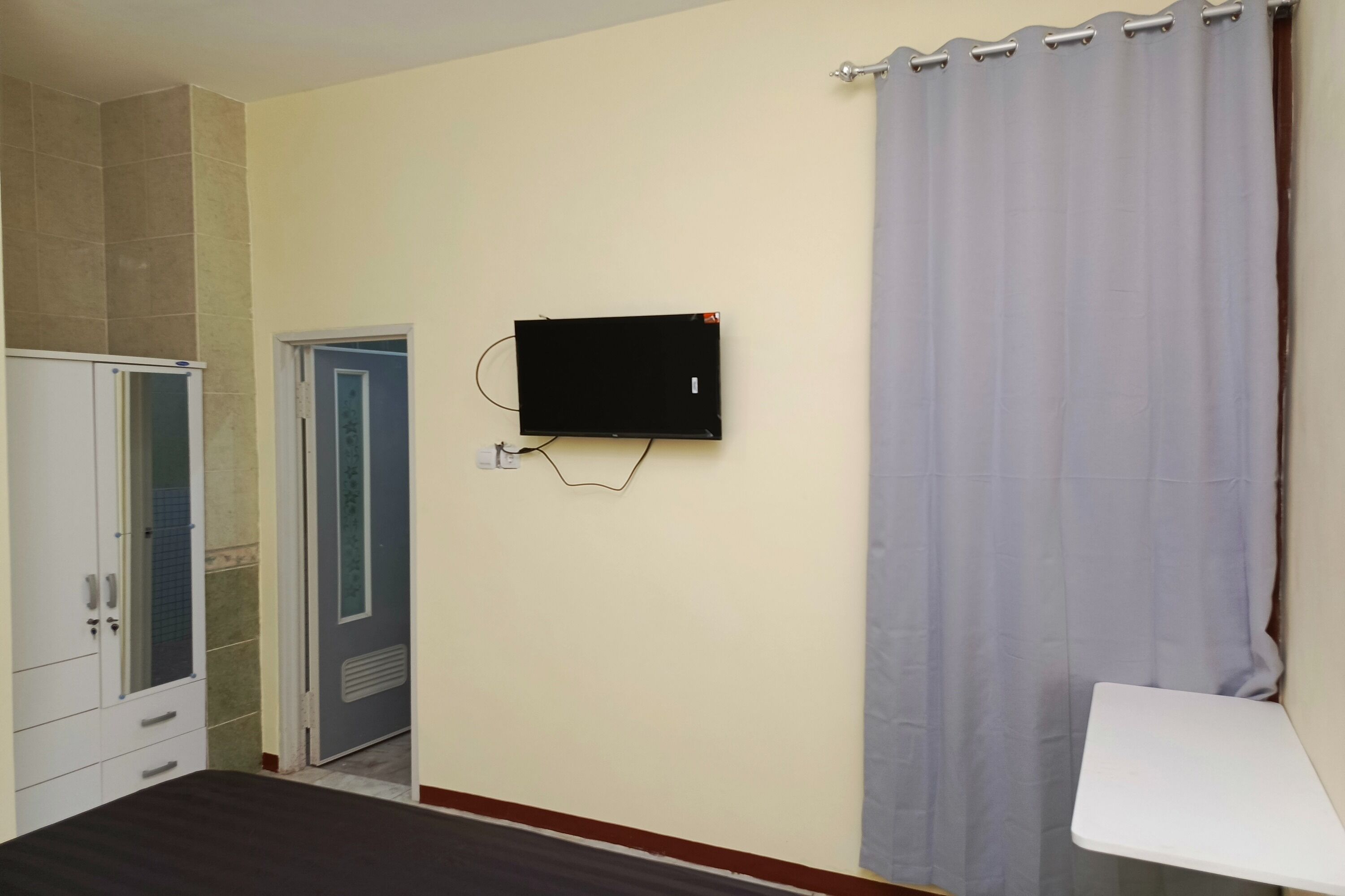 A room with a bed, a TV mounted on the wall, a door, and a curtain.