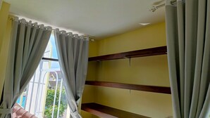 A room with a yellow wall, a window with grey curtains, and wooden shelves. - Bebek Kuning Residence (Malang)