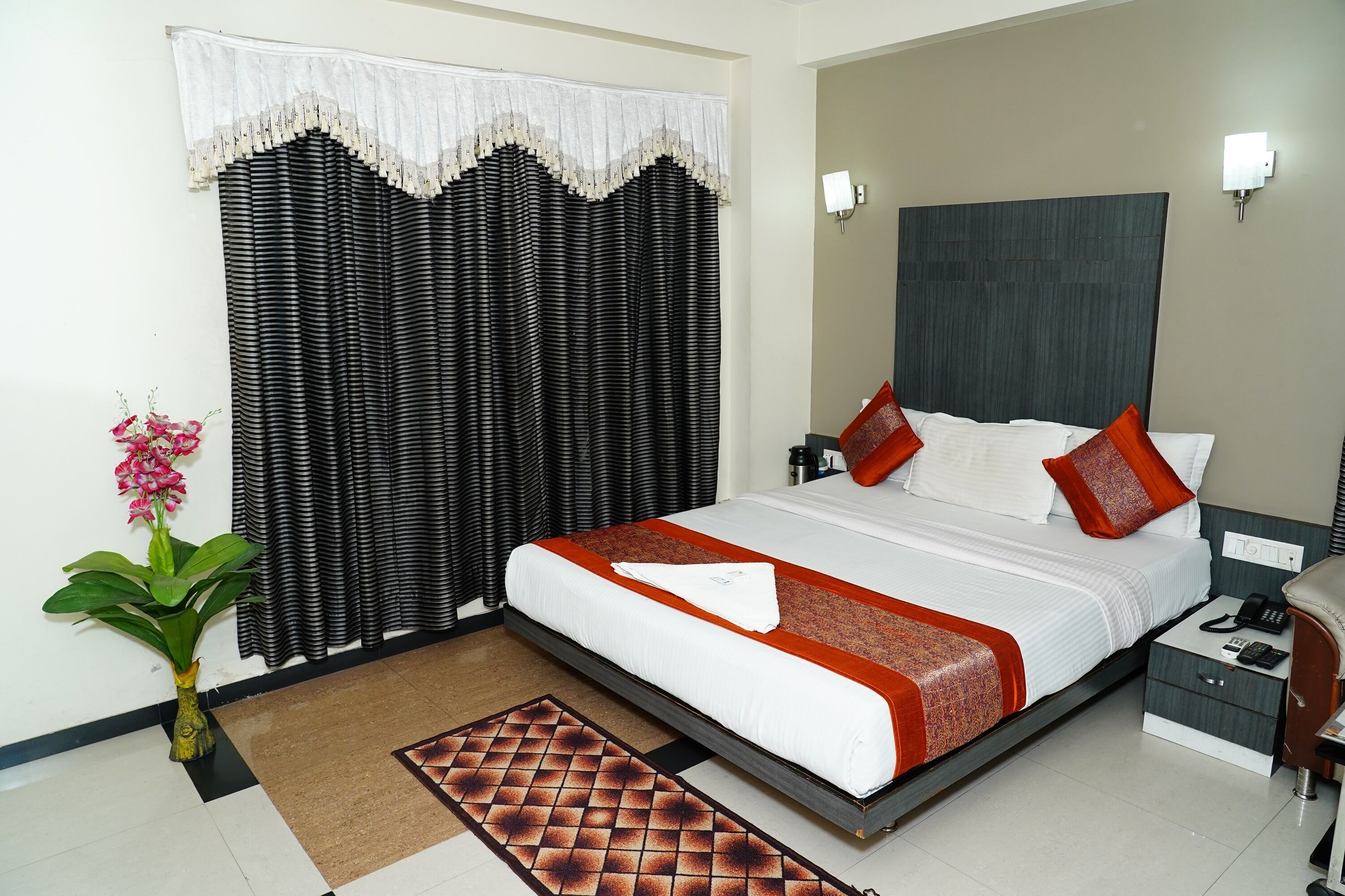 A hotel room with a large bed, a nightstand, a lamp, a potted plant, and a window with curtains.