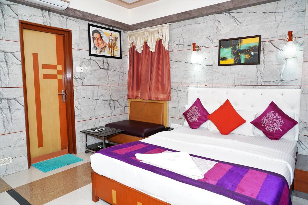 Ein Hotelzimmer mit Marmorwand, einem Bett mit violett-weißer Bettdecke, einer Holztür, einem kleinen Tisch mit einem Laptop und einem roten Vorhang.