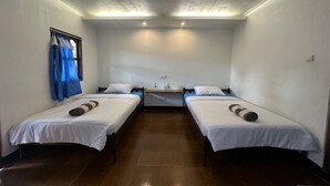 A room with two single beds, a small desk with a chair, a window with blue curtains, and a ceiling light fixture.
