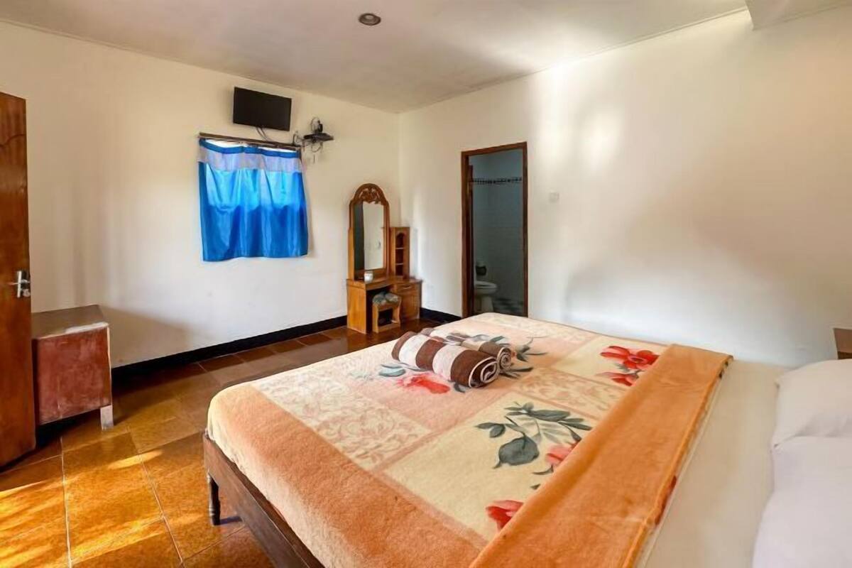Ein Schlafzimmer mit einem Bett, einem Holzschrank, einem kleinen Tisch, einem Spiegel und einem Fenster mit Vorhängen.