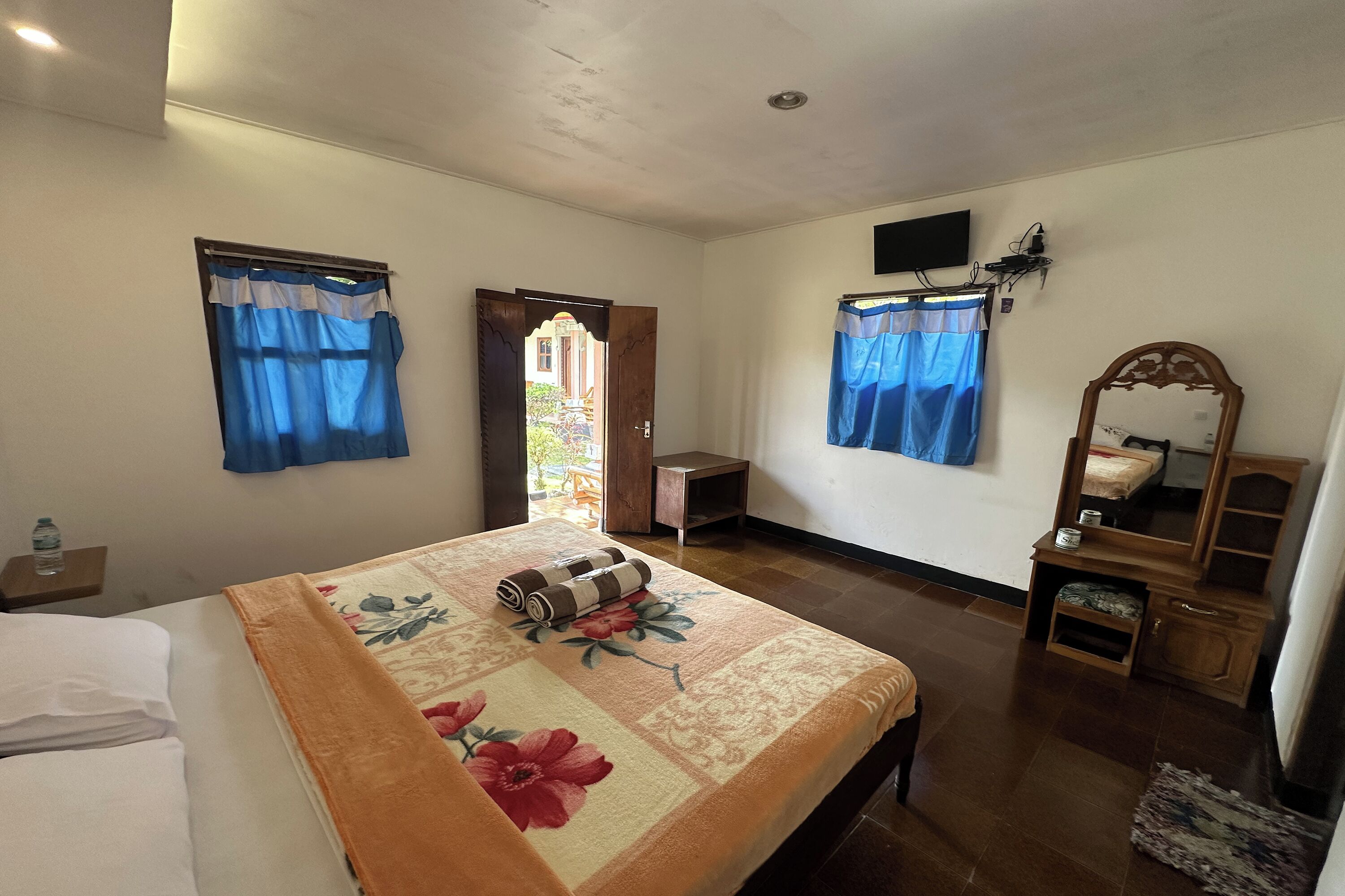 Una habitación de hotel con cama, mesita de noche de madera, espejo, pequeña mesa y puerta que da a un balcón.