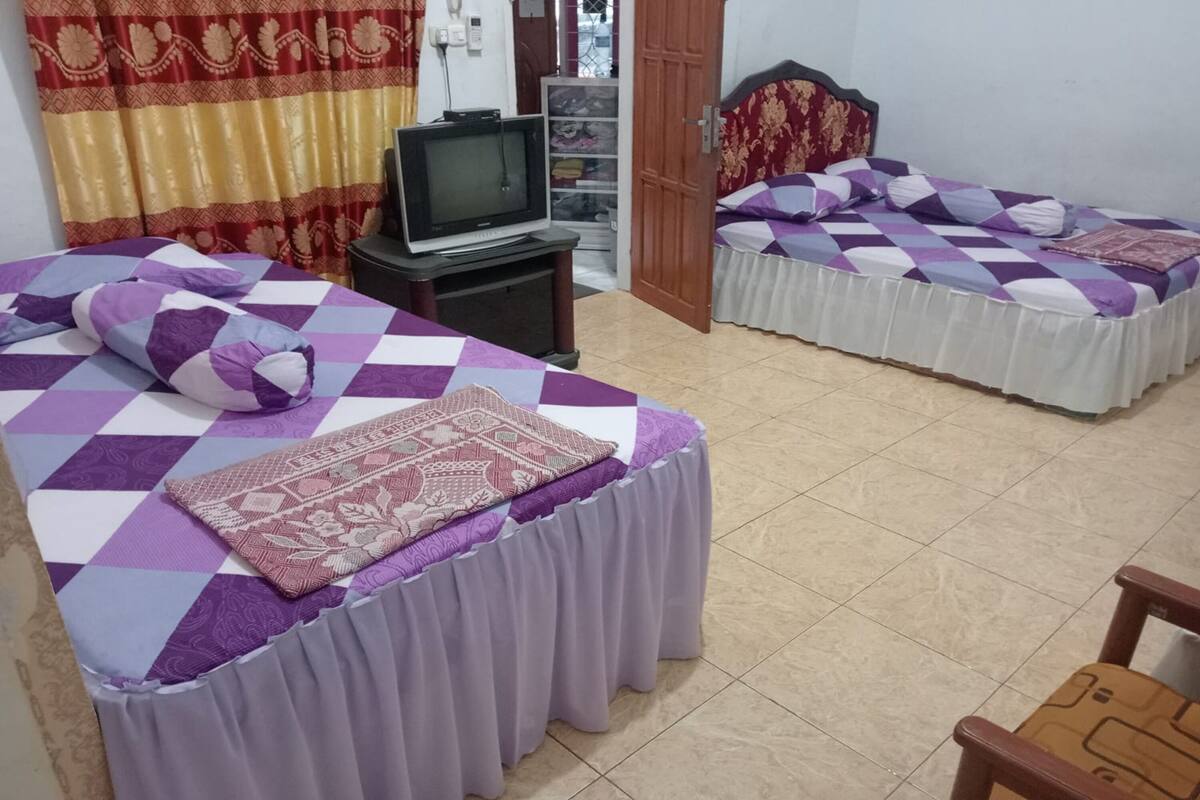 Ein Hotelzimmer mit zwei Betten, jeweils mit einer violett-weißen Karomusterausstattung und passenden Kissen. Zwischen den Betten steht ein kleiner Tisch mit einem Fernseher darauf.