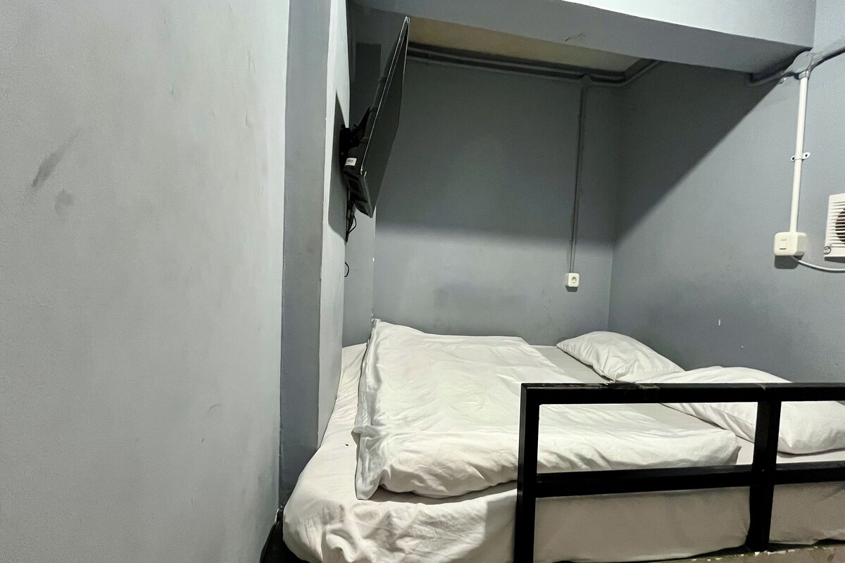 A small, dimly lit room with a single bed, a hanging bag, and a wall-mounted air conditioning unit.