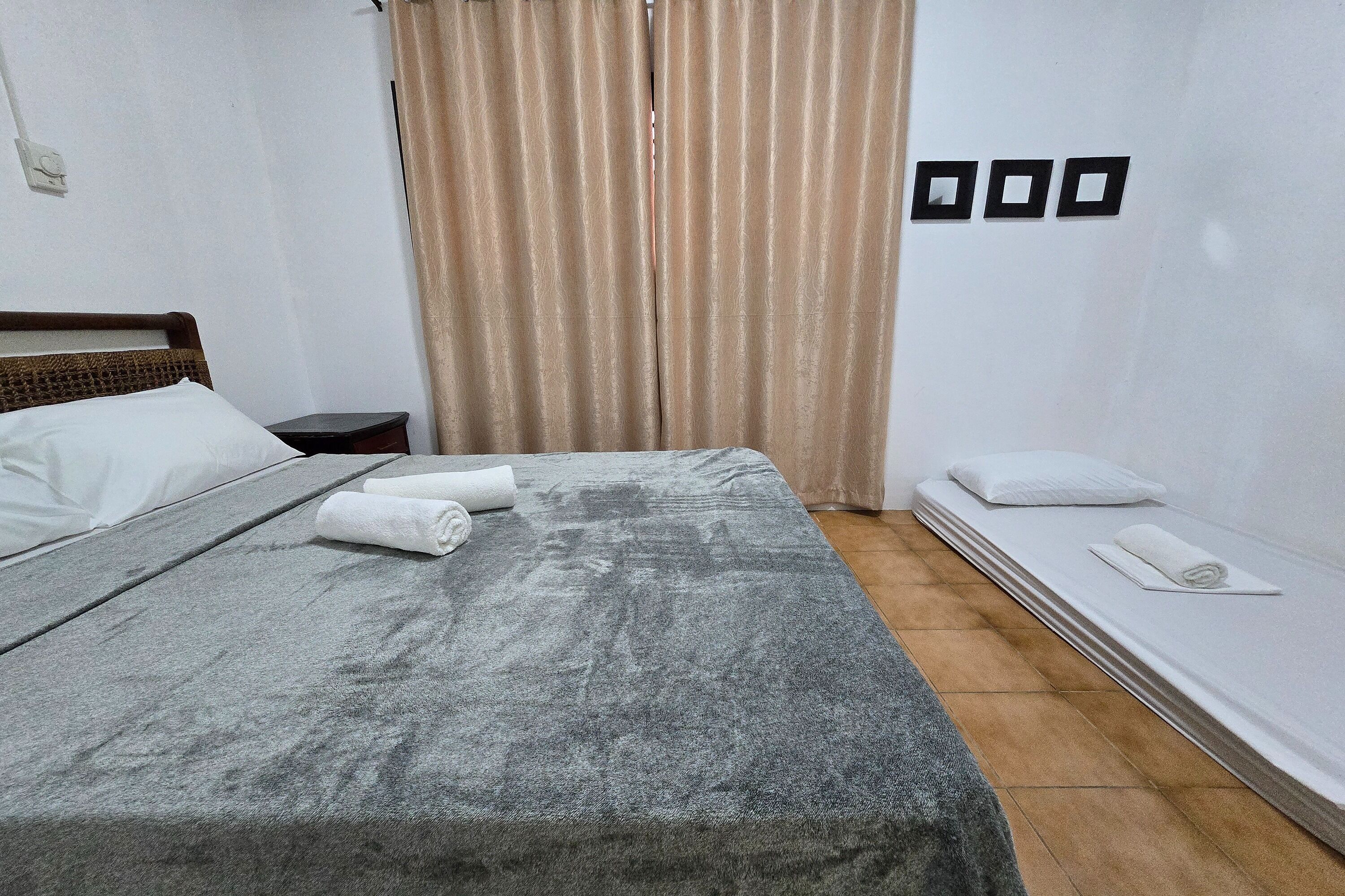 A neatly made bed with a grey blanket and white pillows, a small bedside table with a lamp, and a window with beige curtains.