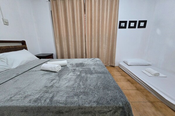 A neatly made bed with a grey blanket and white pillows, a small bedside table with a lamp, and a window with beige curtains. - Grand Villa Laguna (Bay)