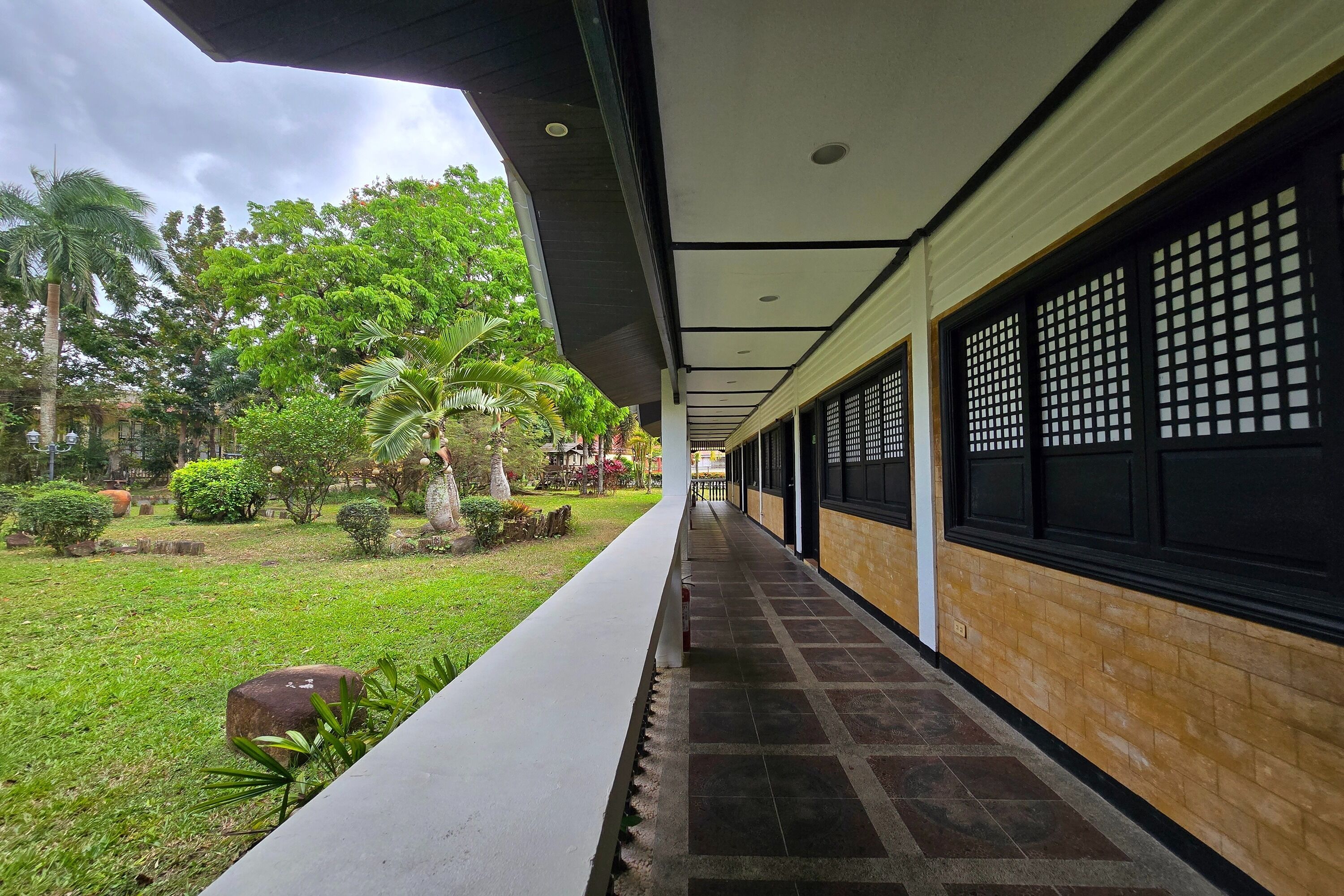 A covered corridor with a tiled floor, a white railing, and a view of a well-maintained garden with trees and shrubs.