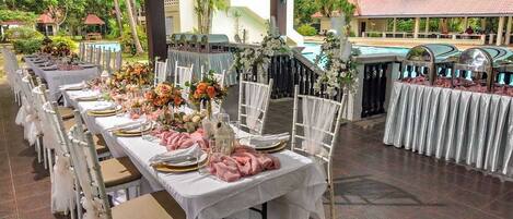 Un salón de banquetes con largas mesas dispuestas para un evento formal, rodeado por una piscina y asientos al aire libre.