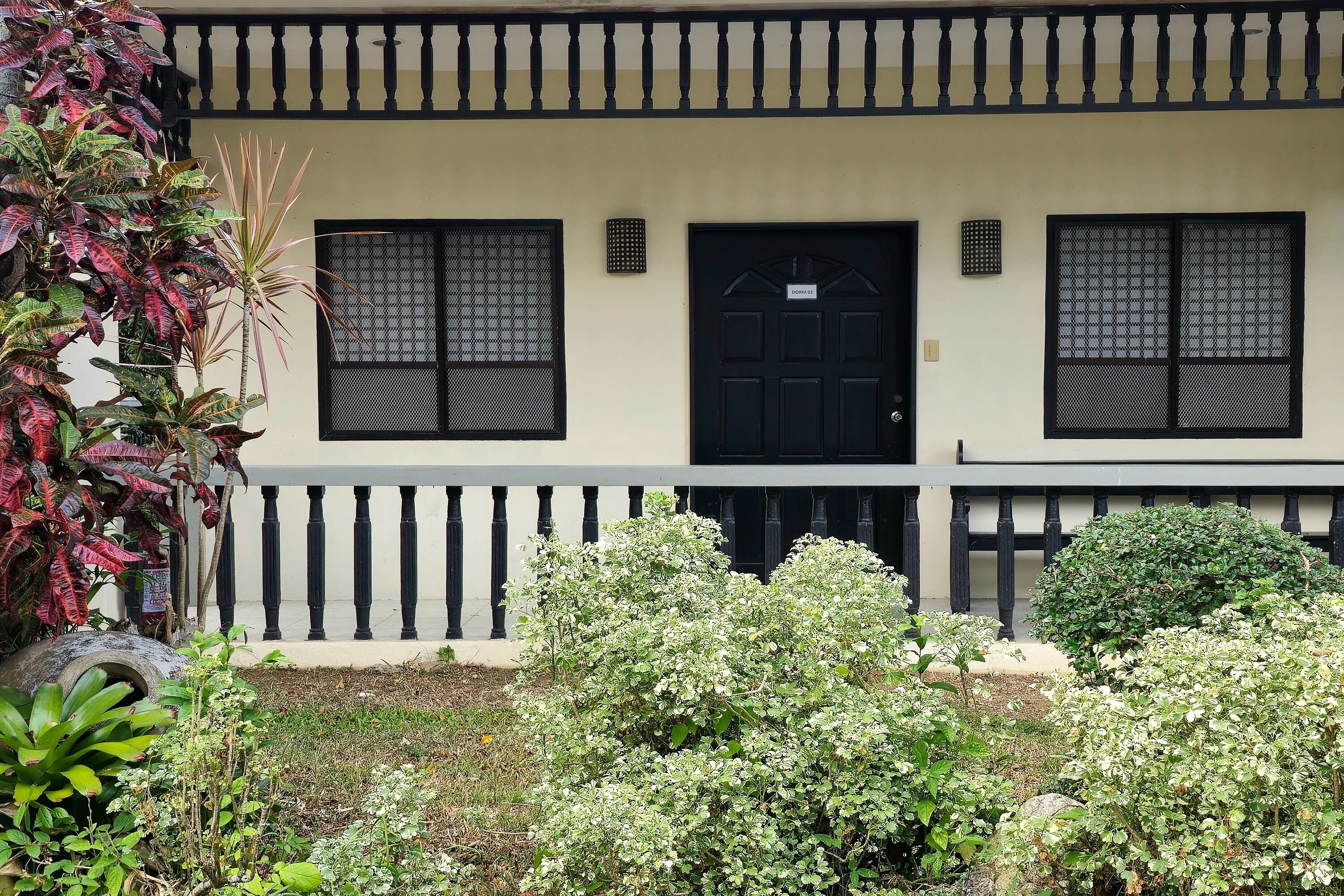 A two-story building with a black door and windows, surrounded by greenery and a white railing.