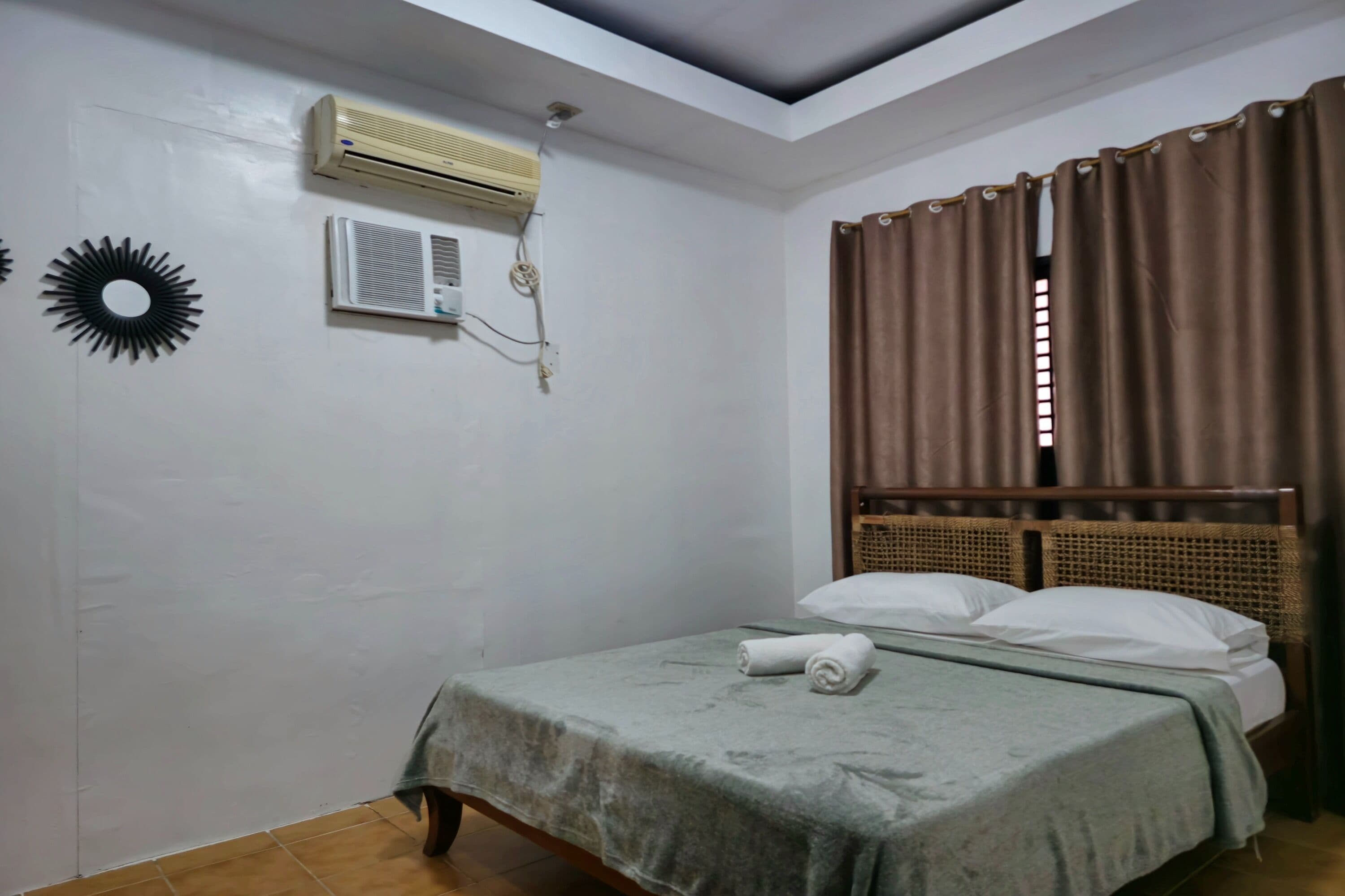 A single bed with a green bedspread, a wall-mounted air conditioner, and a window with brown curtains.
