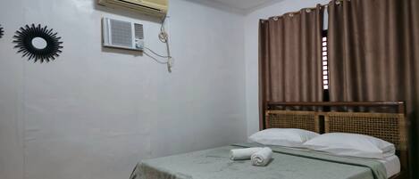 A single bed with a green bedspread, a wall-mounted air conditioner, and a window with brown curtains.