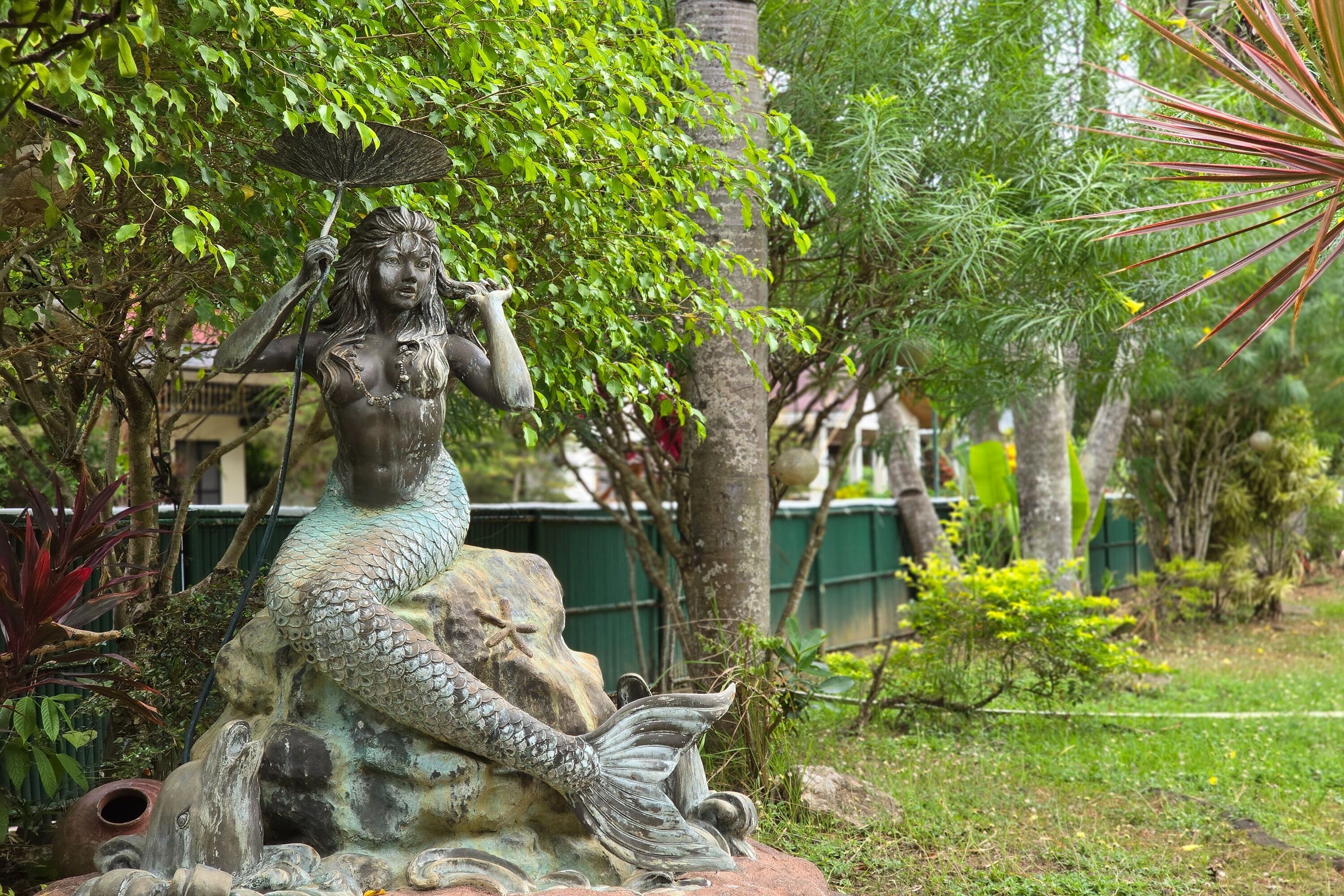 A statue of a mermaid sitting on a rock surrounded by lush greenery.