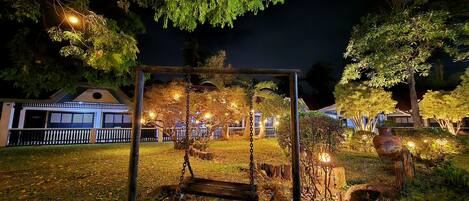 A night scene with a swing set under a tree, surrounded by well-lit garden lights and a fence.