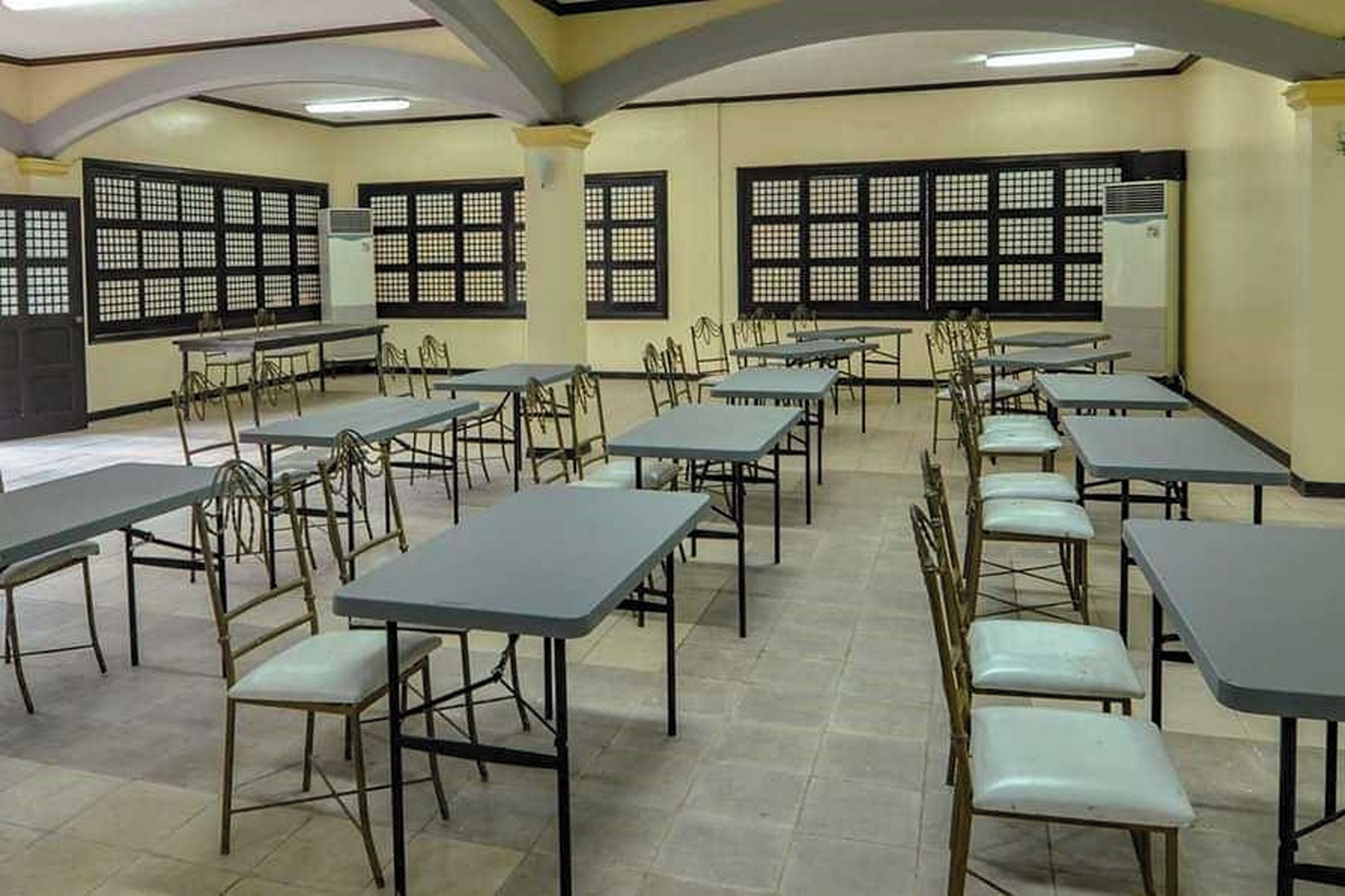 A classroom with rows of tables and chairs, large windows with black frames, and an air conditioning unit on the wall.