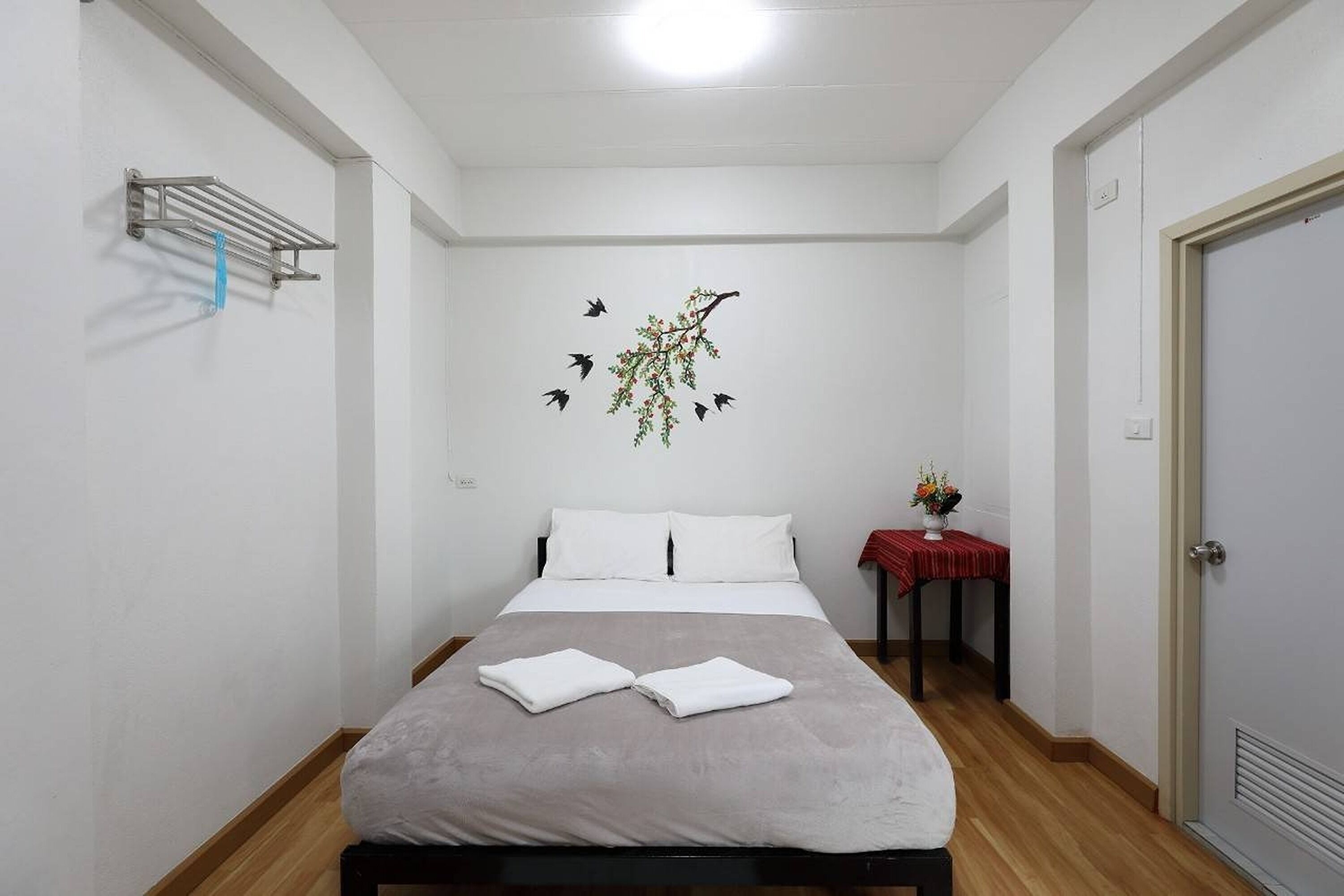 A neatly made bed with a grey blanket and two folded white towels, a wall-mounted shelf with a towel, a decorative branch with birds, and a small table with a vase of flowers.